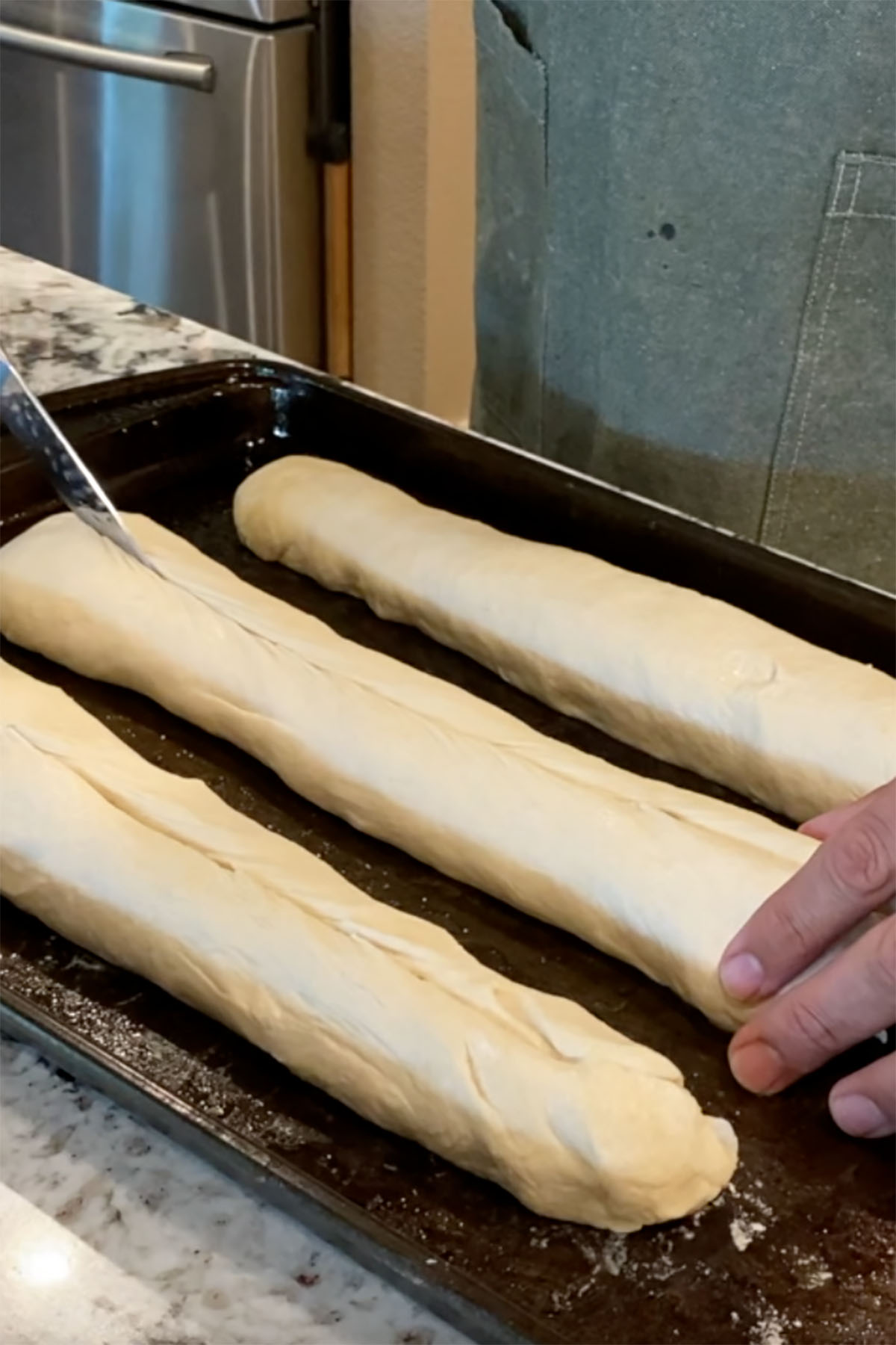 Three loaves of Cuban bread on a baking sheet being scored with a knife.