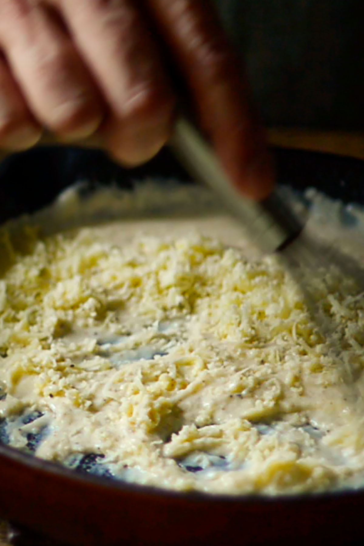 Freshly shredded cheese being folded into Mornay sauce in a cast iron skillet.