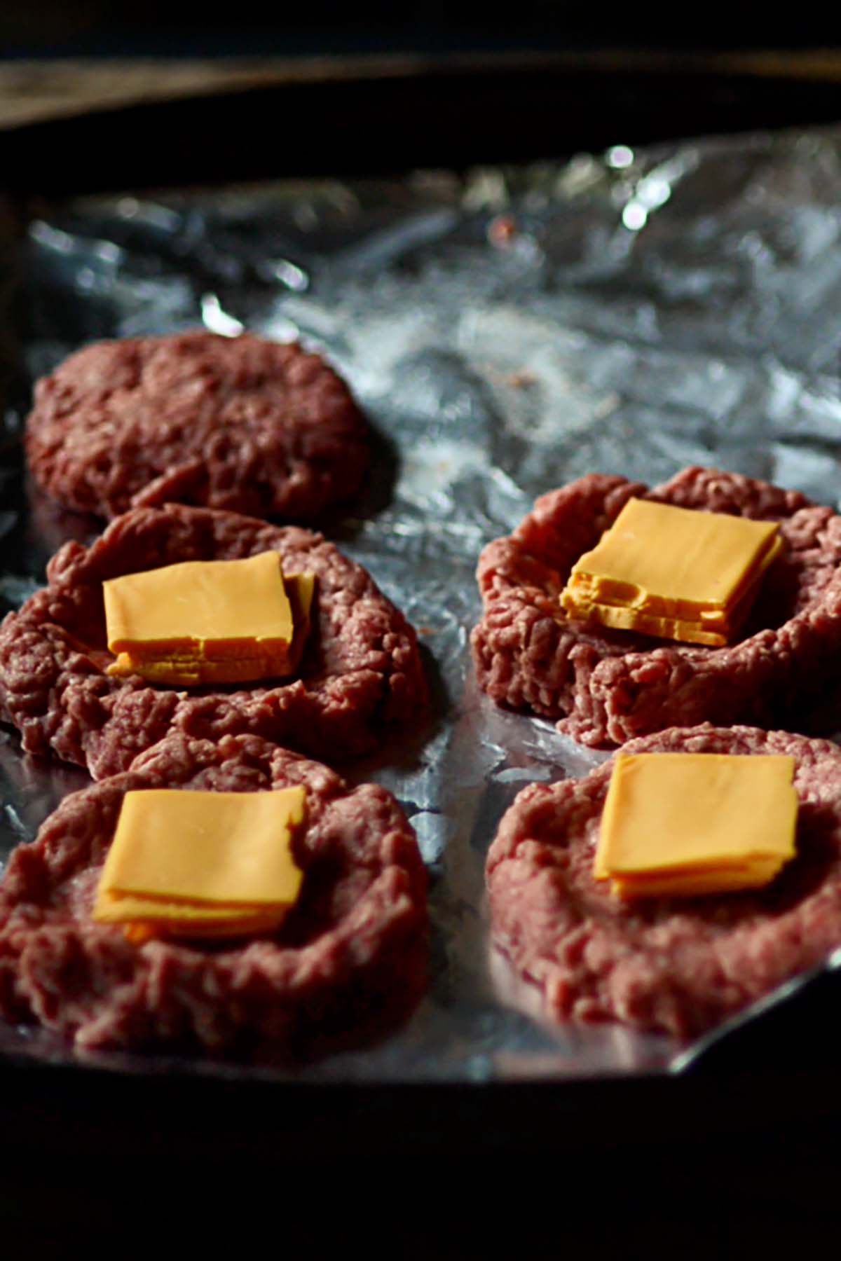 Burger patties on a foil lined cookie sheet with folded slices of cheese on top.