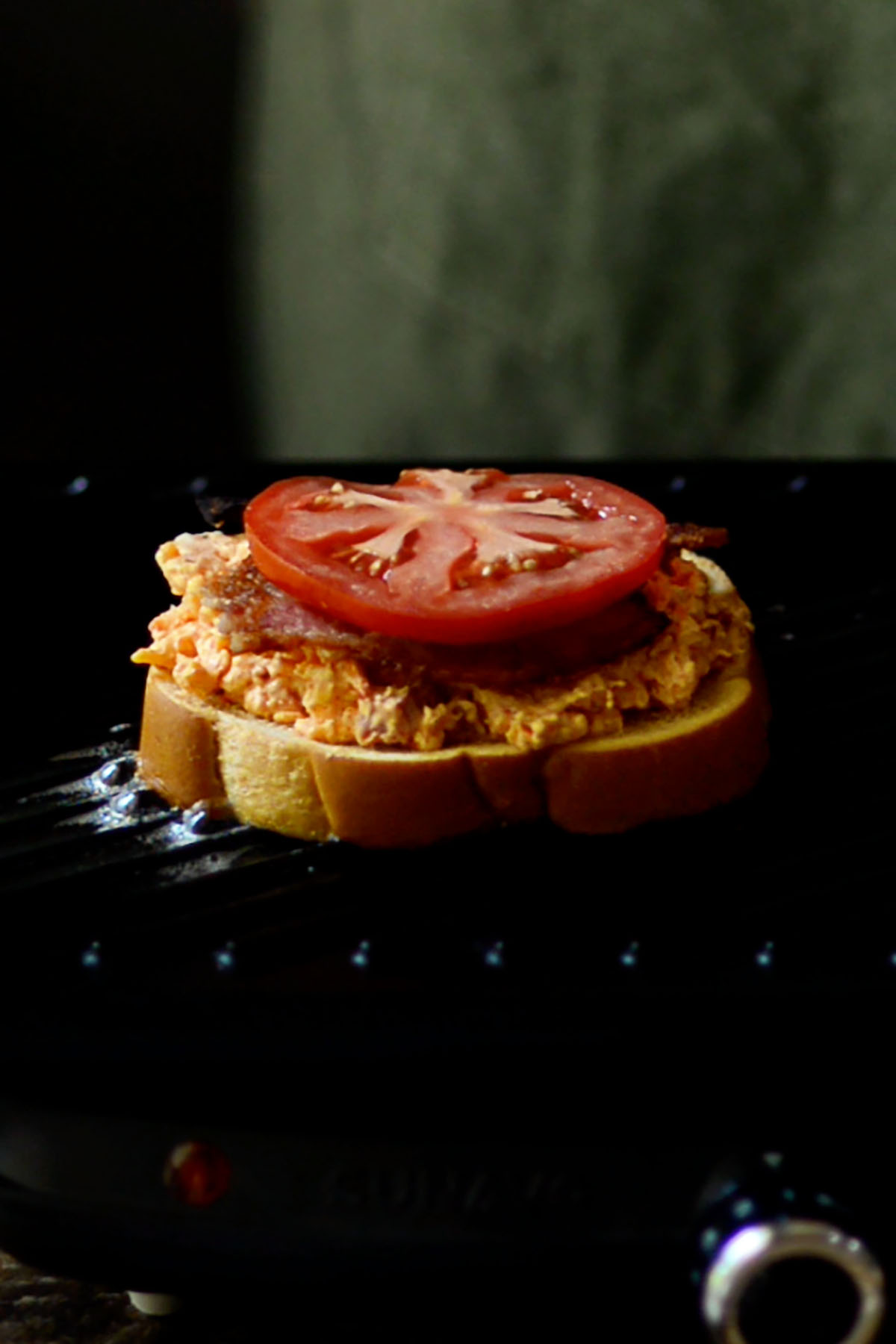 Pimento cheese spread over one slice of Texas toast on a cast iron griddle and topped with a slice of cooked bacon and a tomato slice.