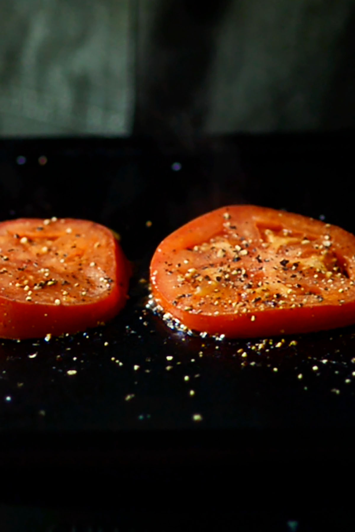 Two sliced tomatoes topped with salt and pepper roasting on a cast iron griddle.