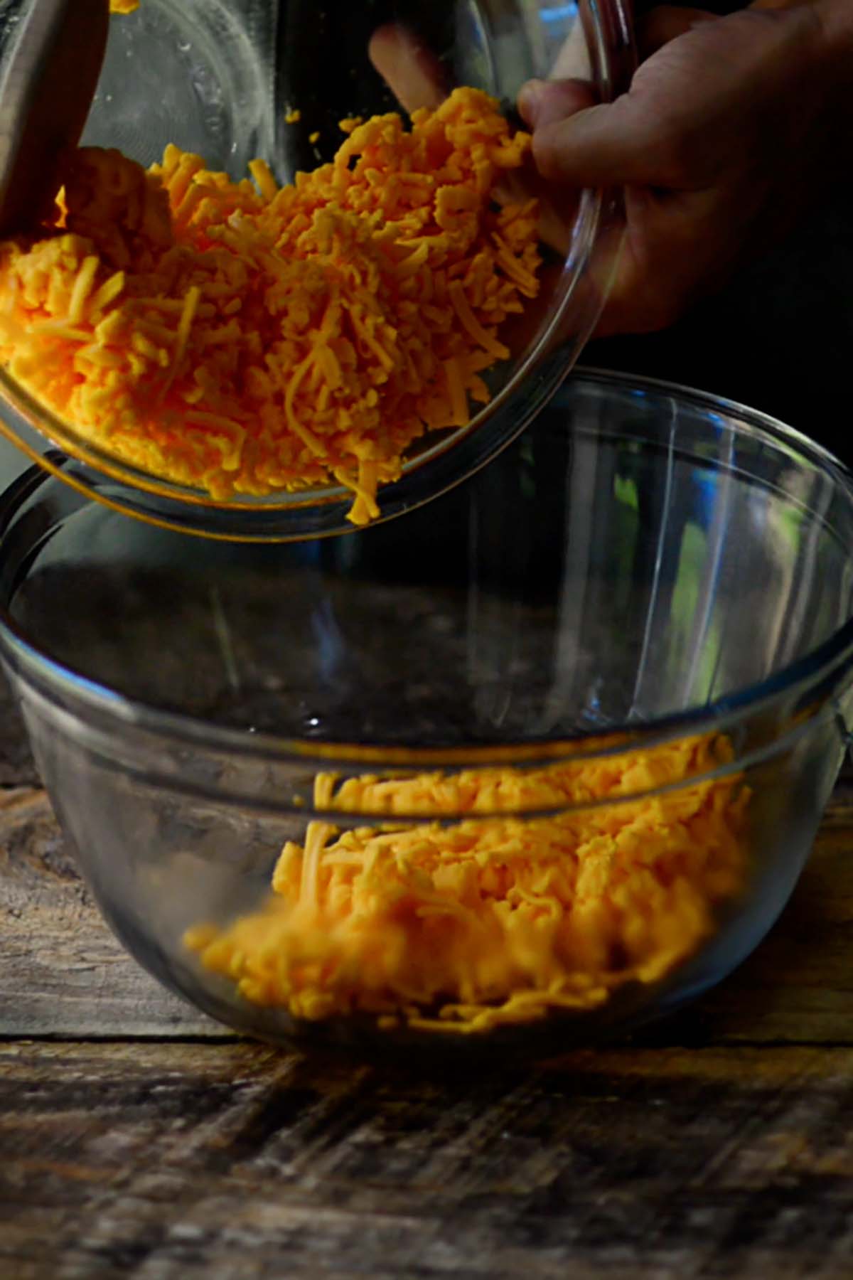 Shredded cheddar cheese being poured into a glass mixing bowl.