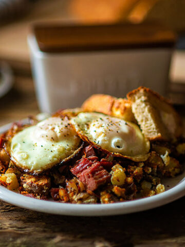 Corned beef hash with two fried eggs and rosemary toast with butter, salt, and bread in the background.