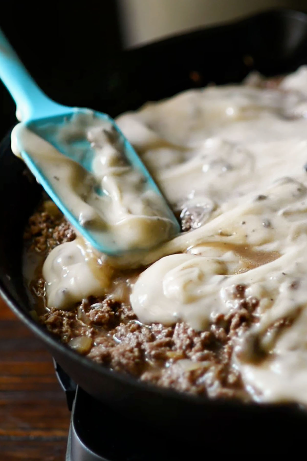Cream of mushroom soup being poured over ground beef and onions in a cast iron skillet.