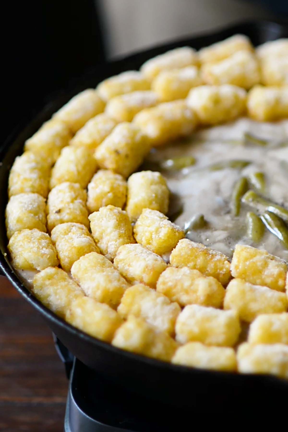Tater tots being placed on top of canned green beans and cream of mushroom soup in a circular pattern around a cast iron skillet.