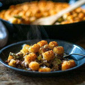Tater Tot Casserole on a blue metal plate with a cast iron skillet full of casserole blurred in the background.