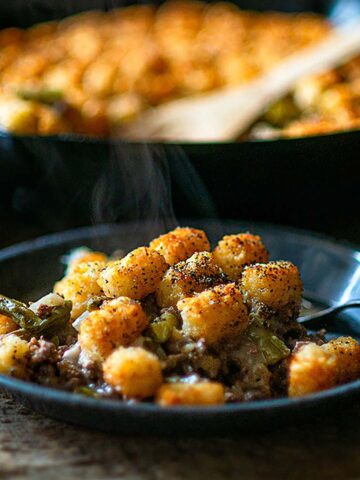 Tater Tot Casserole on a blue metal plate with a cast iron skillet full of casserole blurred in the background.