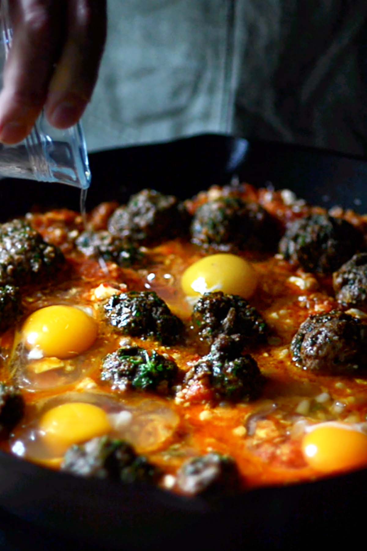 Eggs added to a cast iron skillet full of shakshuka and meatballs.