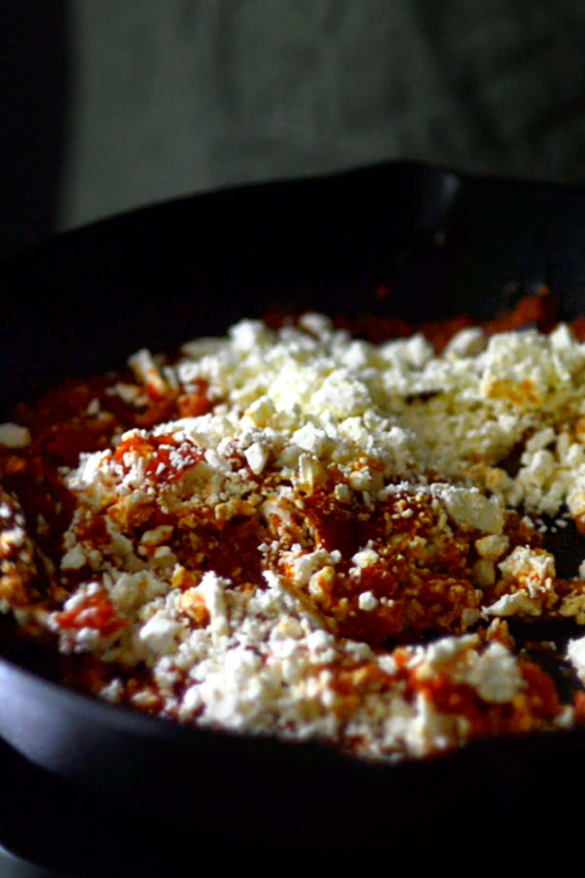 Feta cheese being added to a cast iron skillet with a shakshuka sauce.