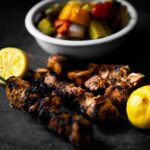 Grilled Mediterranean Salmon Skewers laid out on etched concrete with halved lemons and a blurry bowl of grilled mixed veggies in the background.