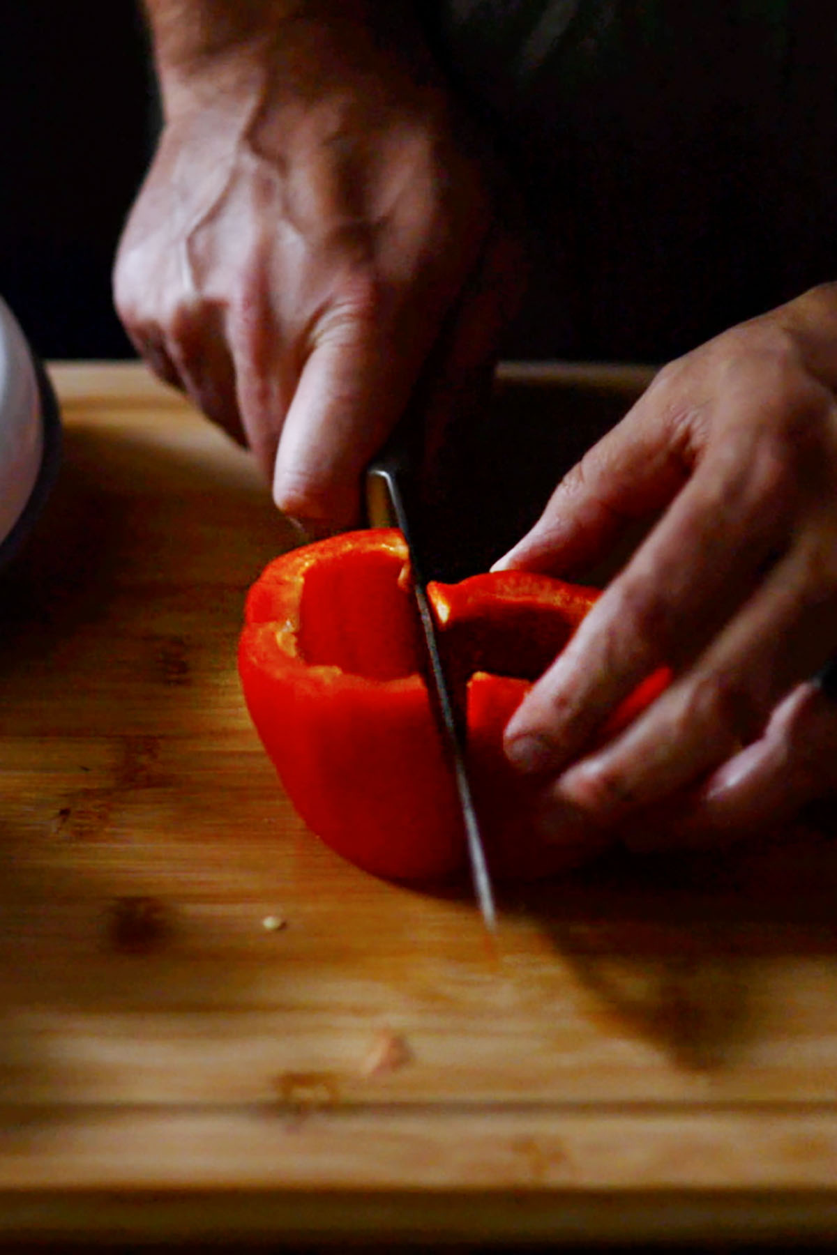 Red bell pepper being sliced in half on a wood cutting board.