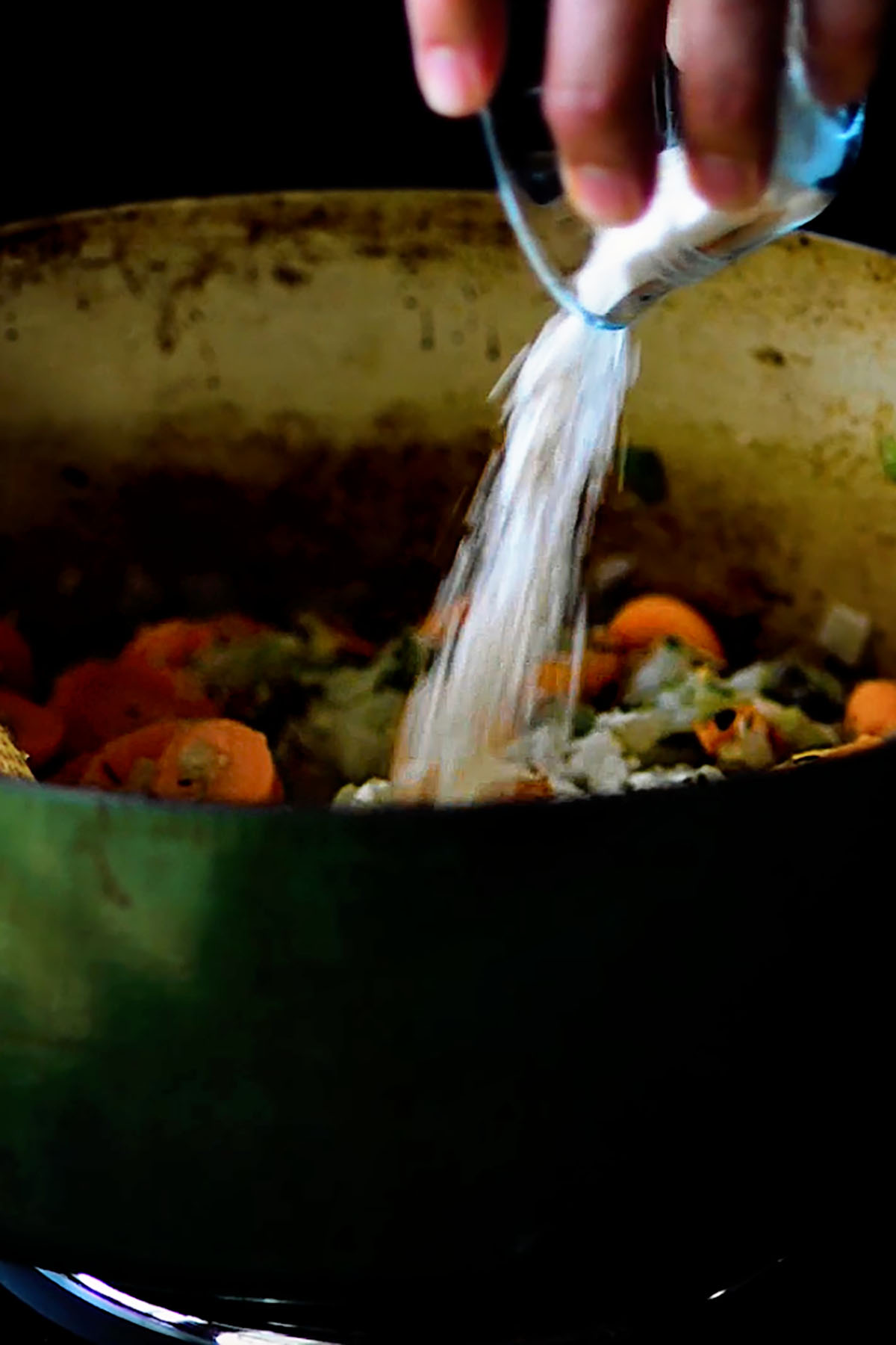 Flour being poured over sautéd vegetables in a dutch oven.