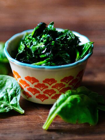 A small red decorated bowl of sautéed spinach with fresh spinach leaves laid next to the bowl.