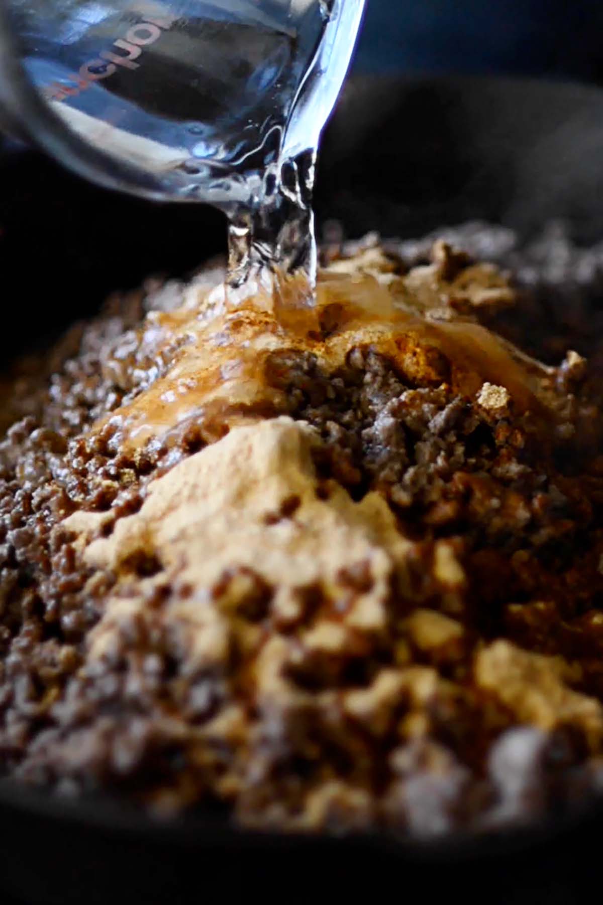 Water being poured into ground beef topped with with taco seasoning in a cast iron skillet.