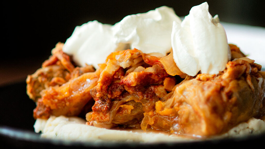 A close-up of a savory dish featuring a generous portion of stuffed cabbage rolls topped with a dollop of sour cream, served on a bed of creamy mashed potatoes. The cabbage rolls are visibly filled with a rich, tomato-based mixture.