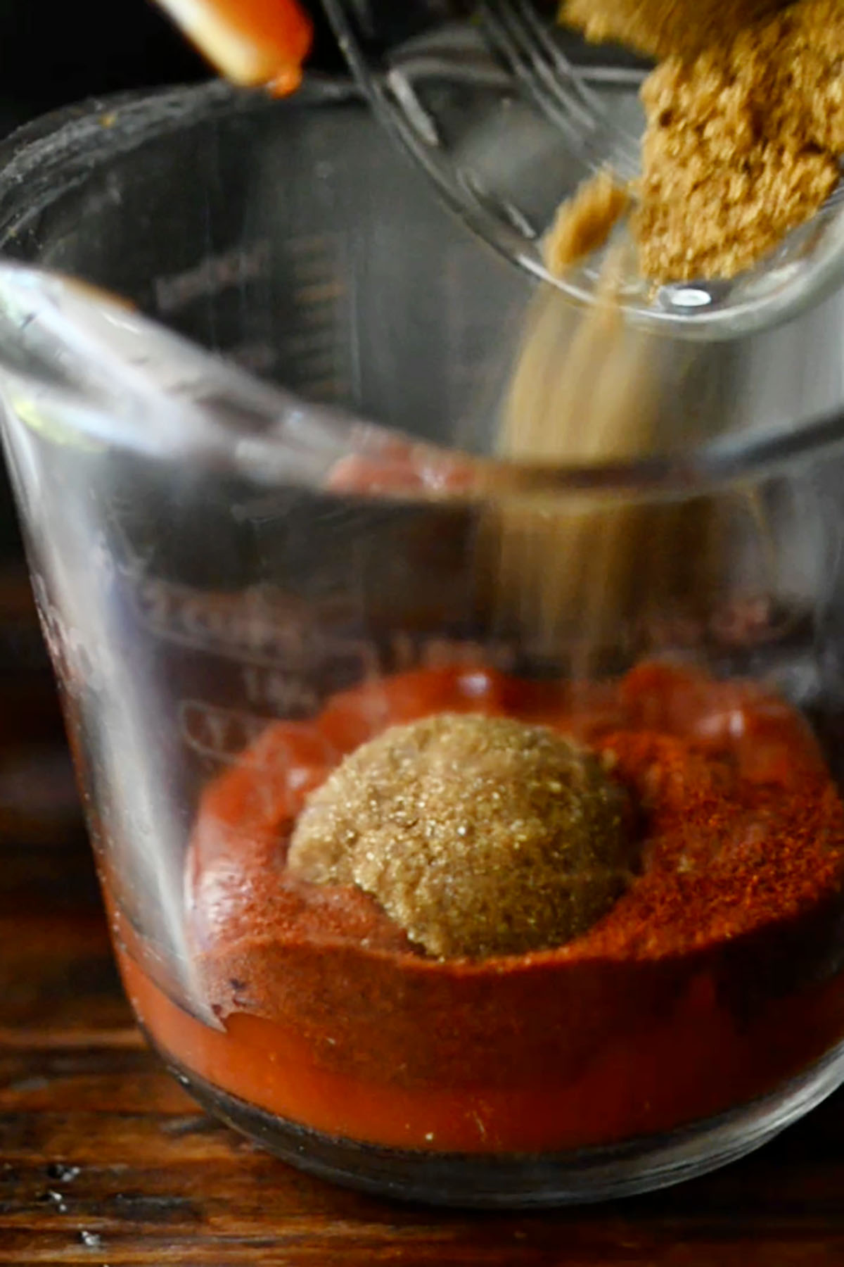 Brown sugar being added a ketchup mixture in a glass measuring cup.