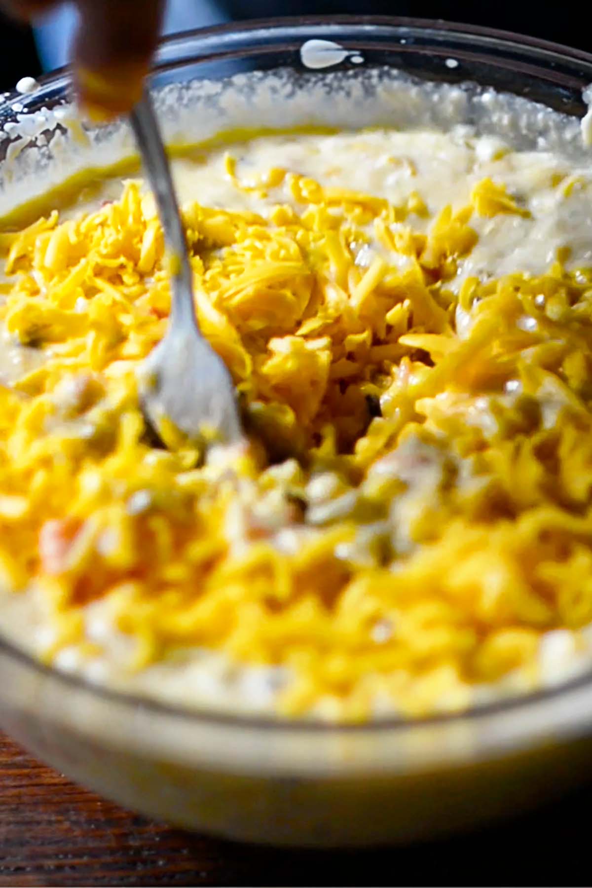 Shredded cheese being added to a corn pudding mixture in a glass mixing bowl.