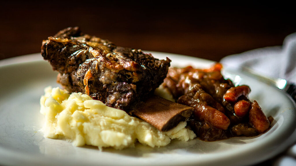 A dinner plate featuring a tender piece of beef short rib resting on a bed of creamy mashed potatoes. A serving of richly seasoned beans with bell peppers is placed beside the meat. The dish is illuminated with warm, inviting lighting.