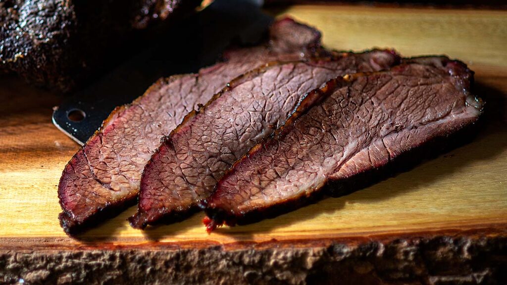 Three slices of smoked brisket with a dark, seasoned crust are laid out on a wooden cutting board. The meat appears tender and juicy with a rich brown color, showcasing visible marbling and a thin smoke ring along the edges.