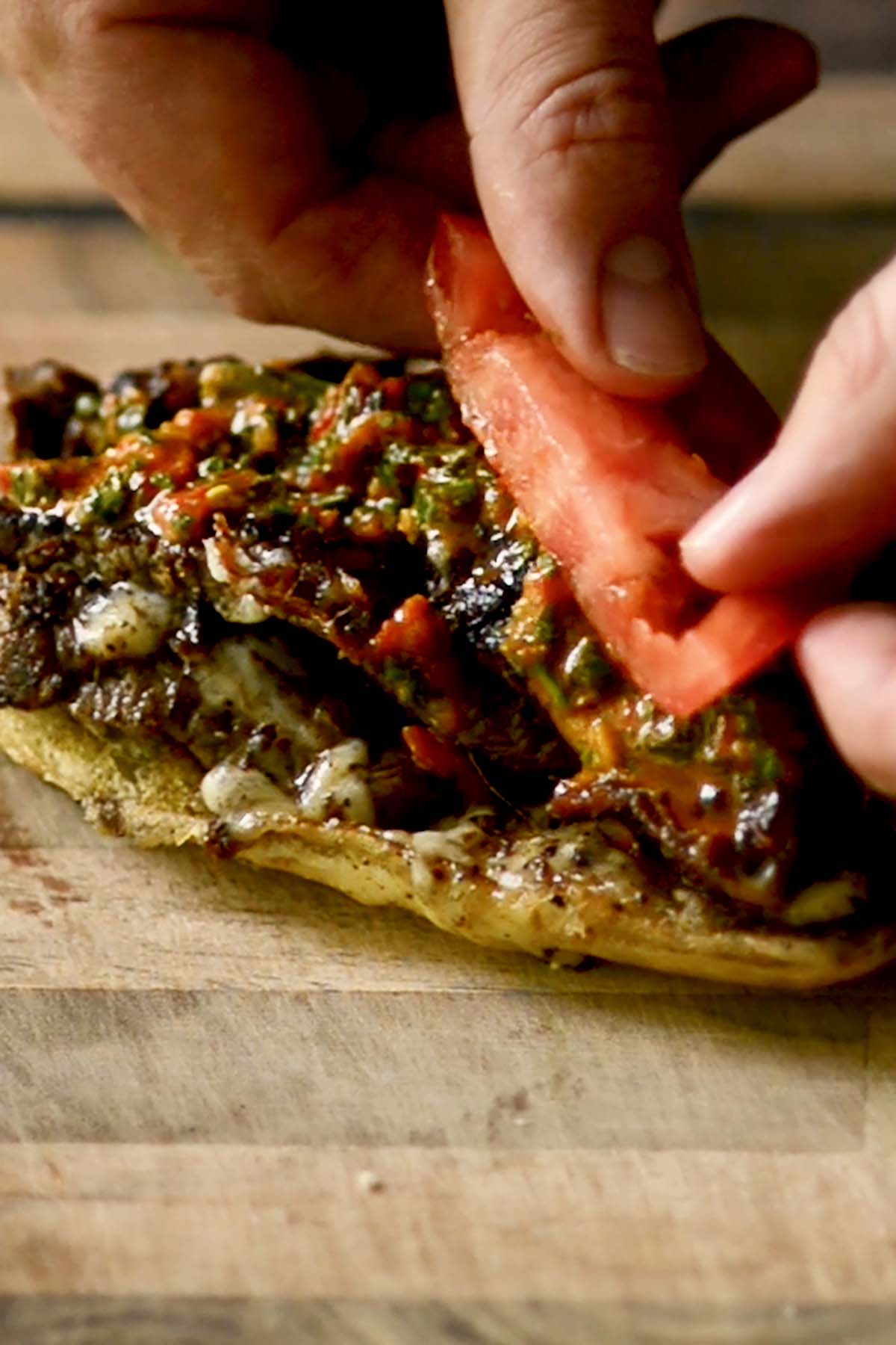 A Churrasco Italiano Steak sandwich being topped with red chimichurri and freshly sliced tomato.