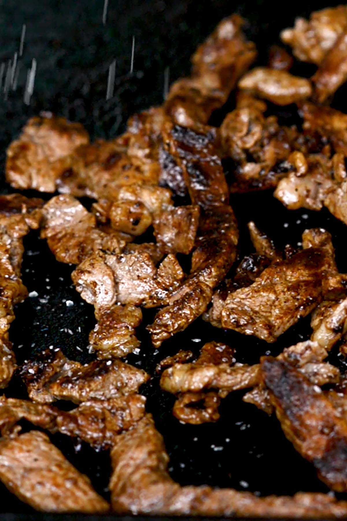 Sliced steak being seasoned with salt and pepper while cooking on a Blackstone griddle.