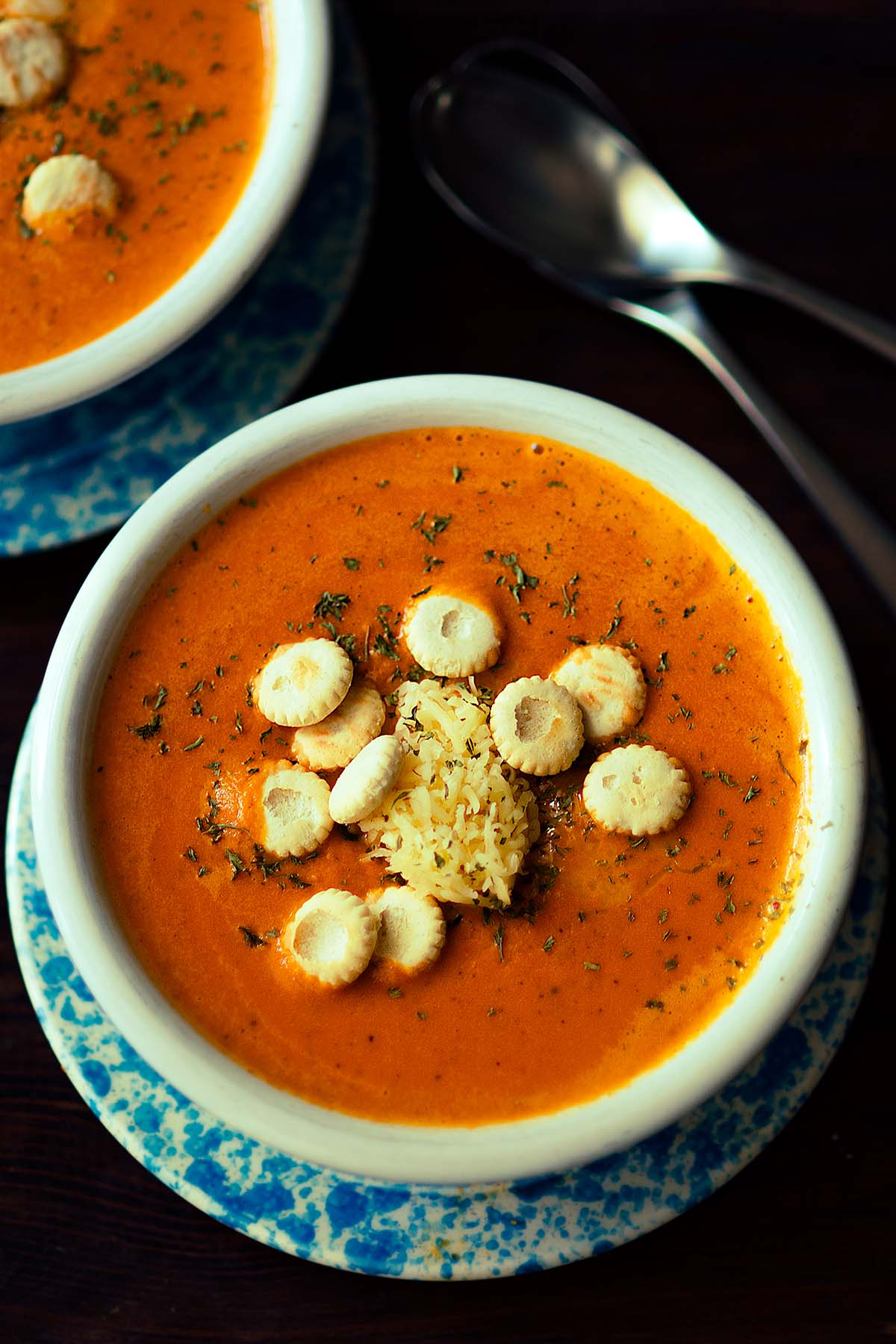 A bowl of roasted red pepper and gouda soup served in a white bowl and topped with oyster crackers, grated gouda, and fresh parsley.