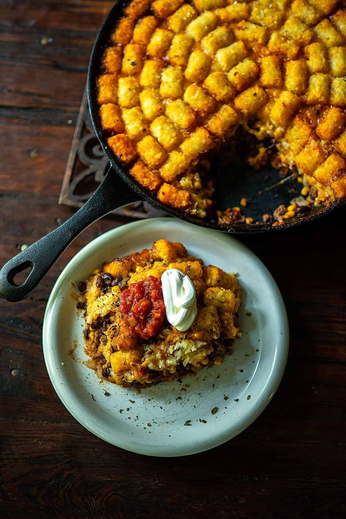 Taco Tater Tot Casserole Served family style in a cast iron skillet and a portion scooped on a plate then topped with salsa, sour cream, and dried Mexican oregano.