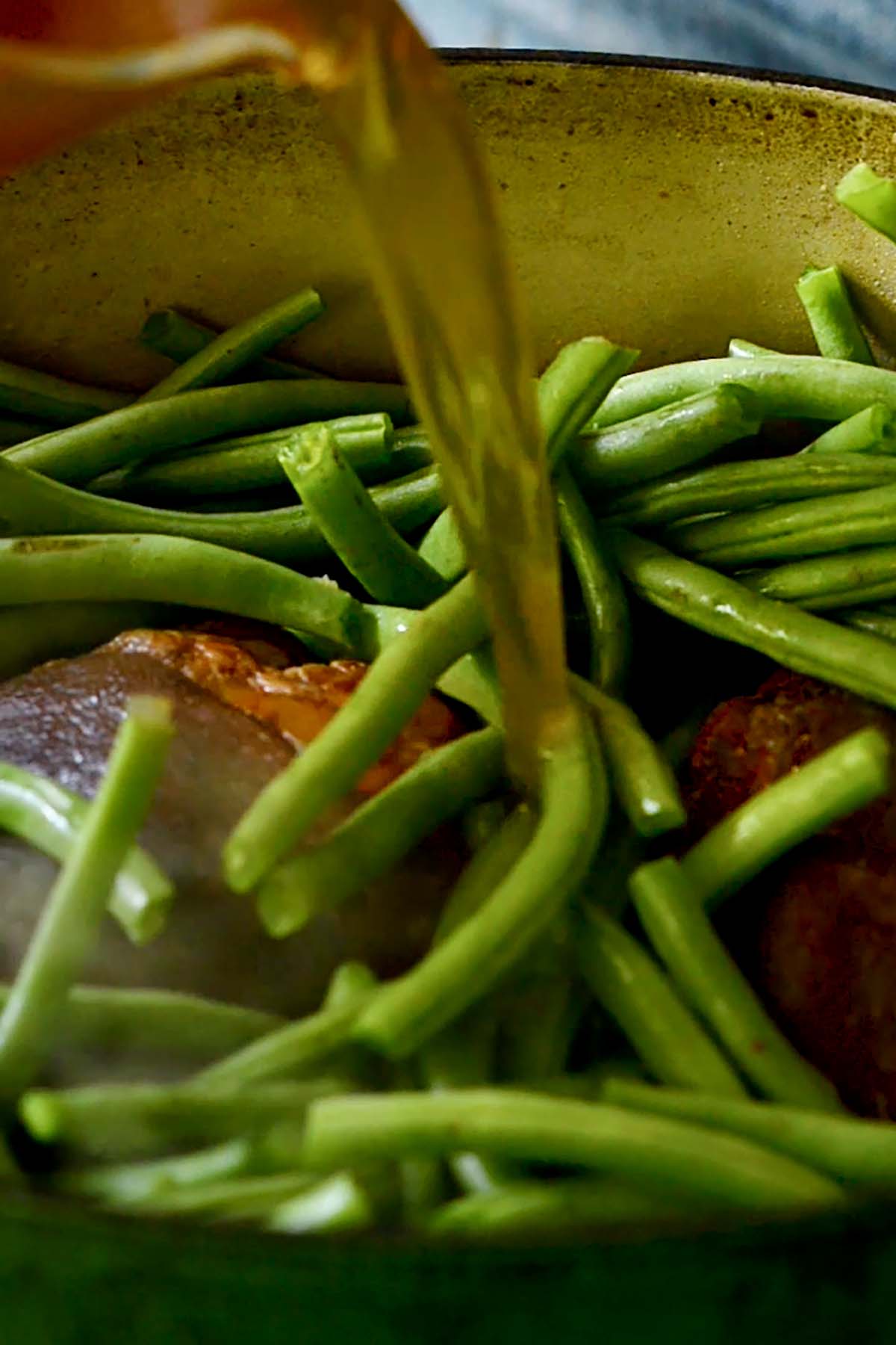 Chicken stock being poured over fresh sliced green beans and ham hocks in a dutch oven.