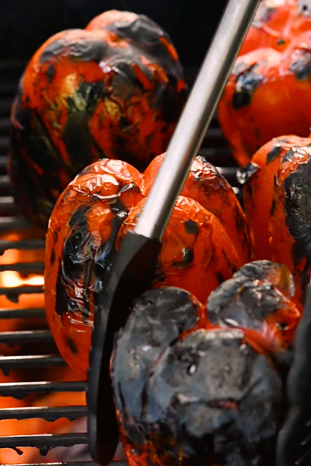 Red bell peppers roasting over an open fire on a charcoal grill.