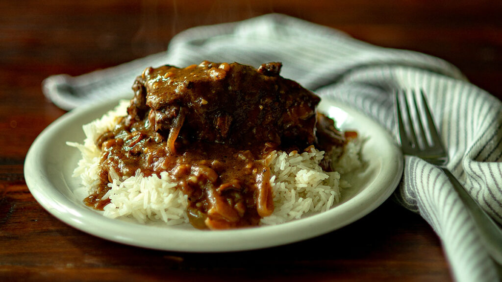 A plate of white rice topped with a generous serving of beef stew, featuring chunks of tender meat and a rich brown gravy with visible slices of onion. The plate is placed on a wooden table next to a striped cloth napkin and a fork. Steam rises from the dish.