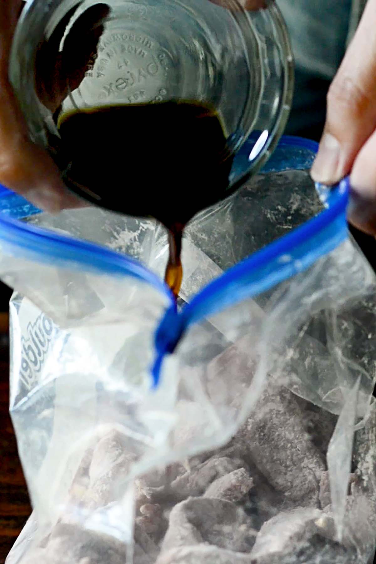 Strips of top sirloin coated in a corn starch in a plastic bag with soy sauce being poured into the bag.