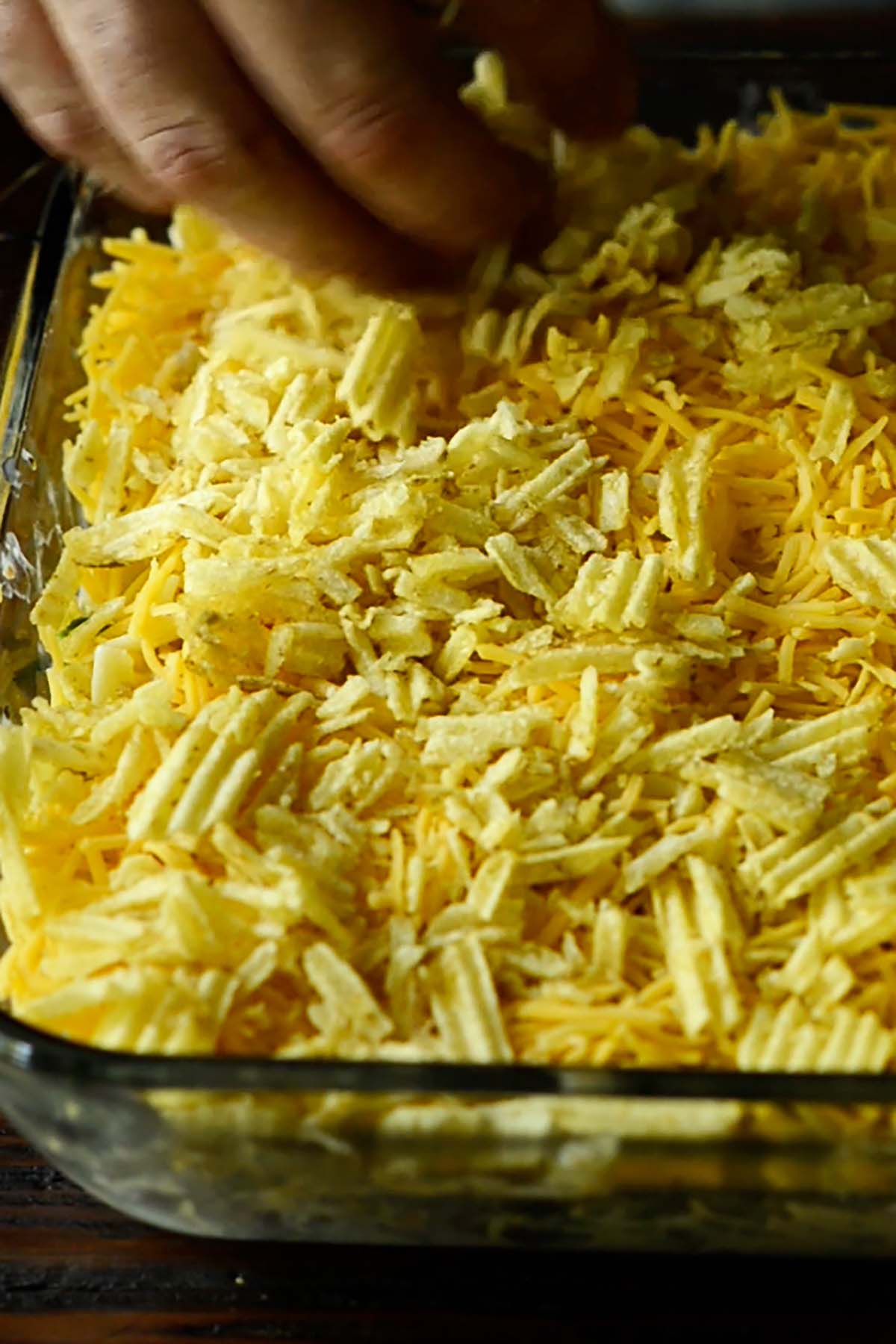 Crunched up potato chips being spread evenly across the top of a cheesy unbaked tuna noodle casserole.