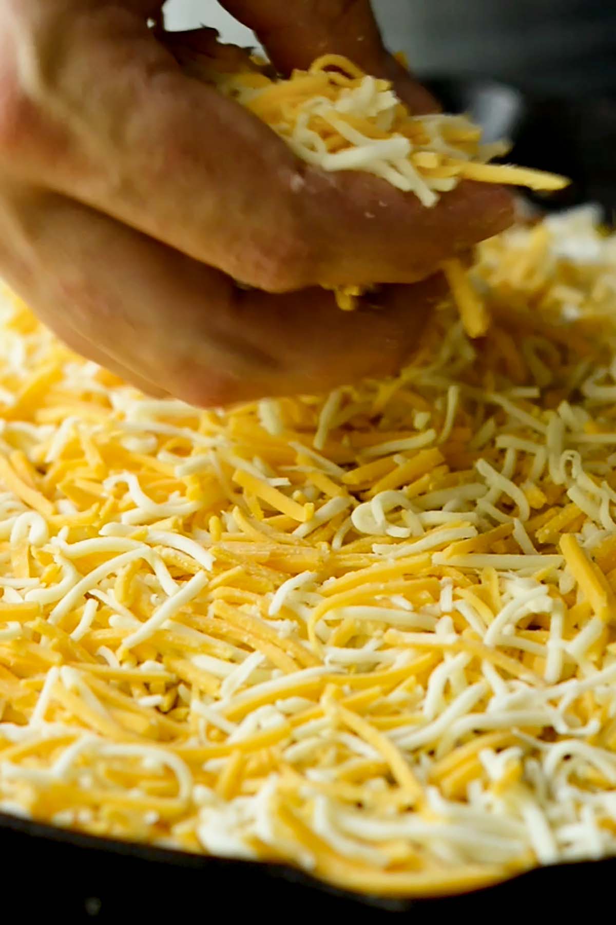 Shredded cheese being evenly spread over a taco tater tot casserole in a cast iron skillet.