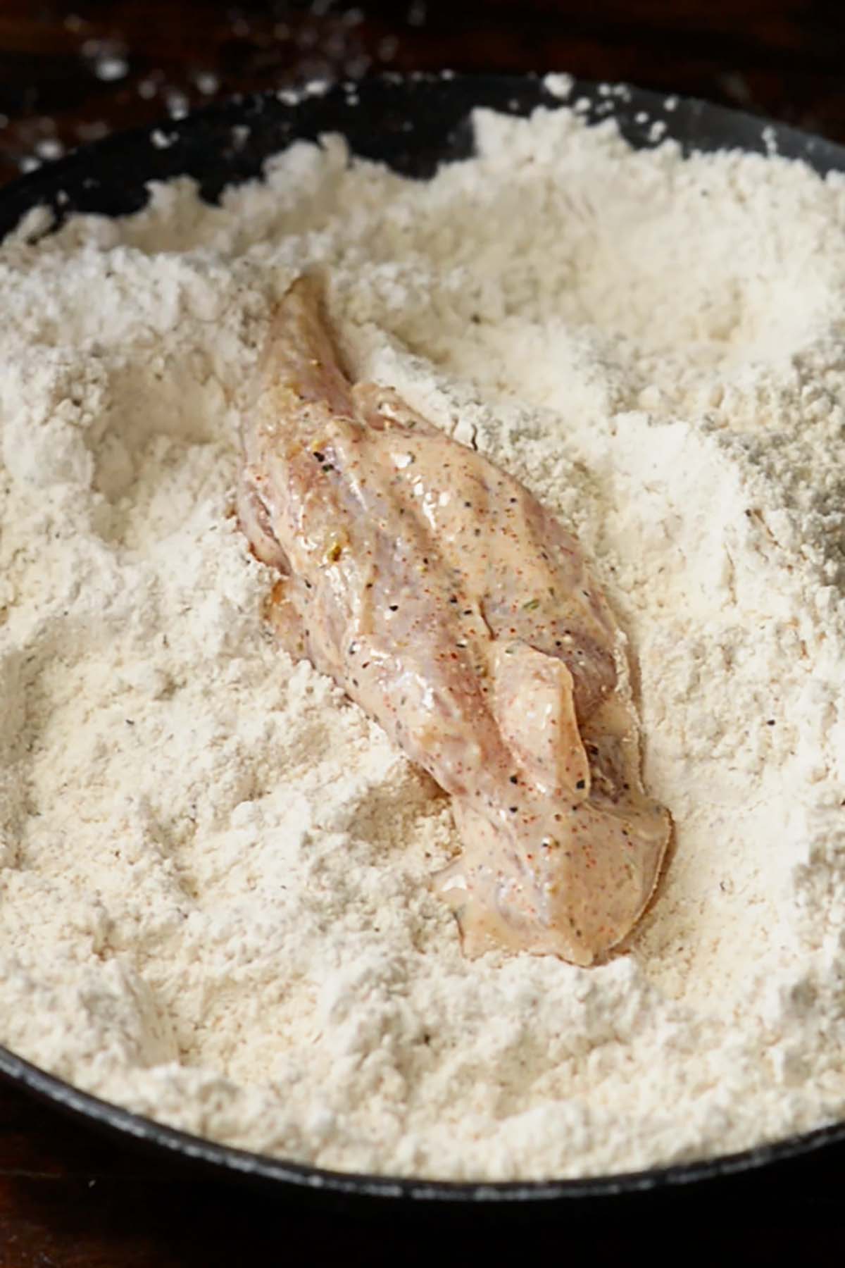 A marinated chicken strip in a shallow dish full of seasoned flour.