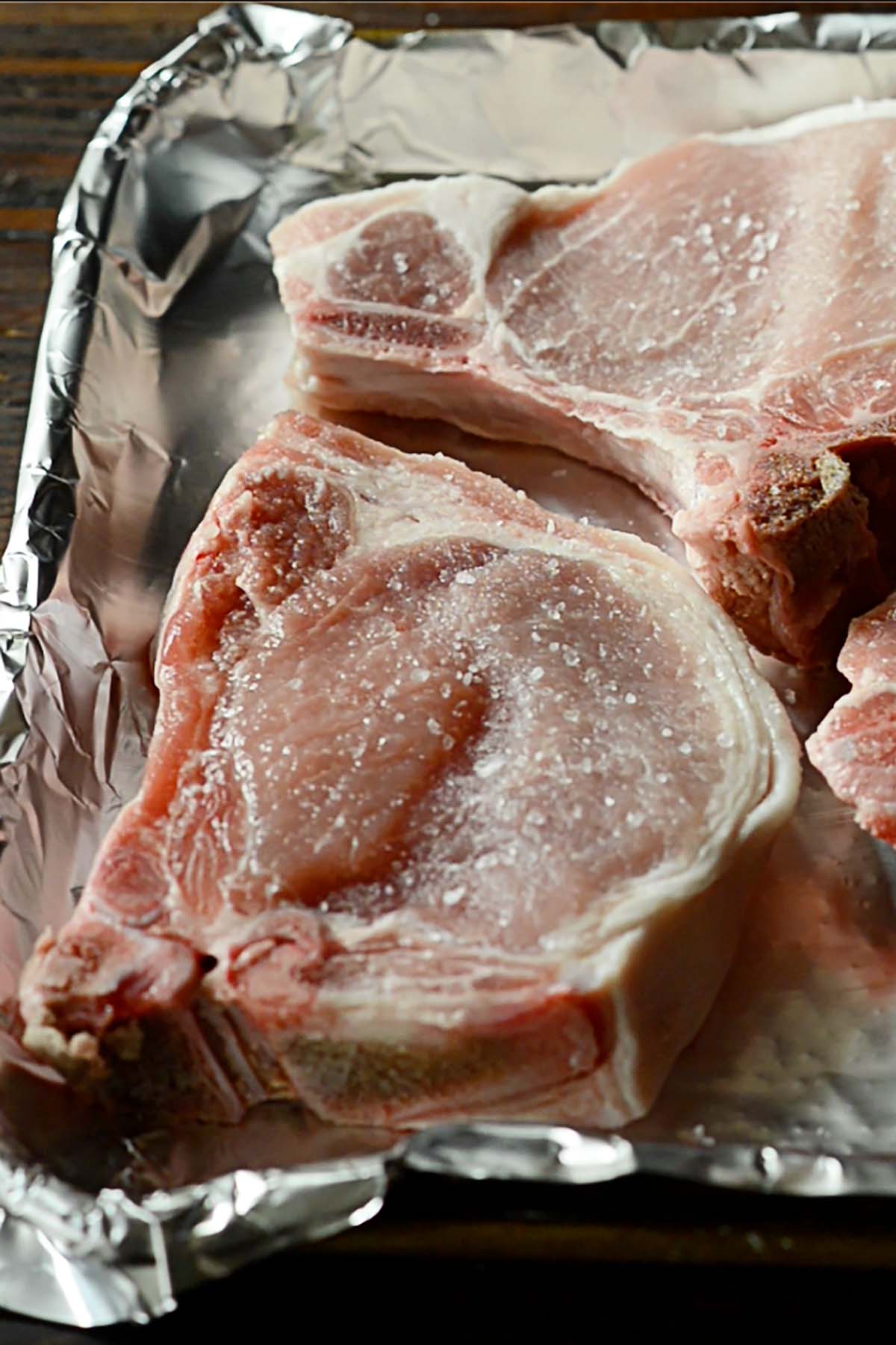 Raw thick cut pork chops on a foil lined tray seasoned with coarse salt.