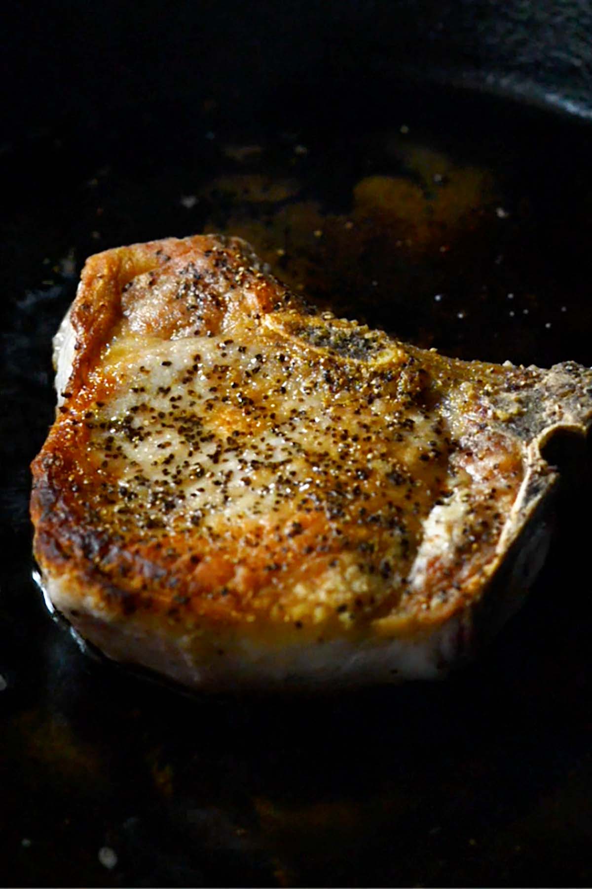 A thick cut pork chop being seared in a cast iron skillet.