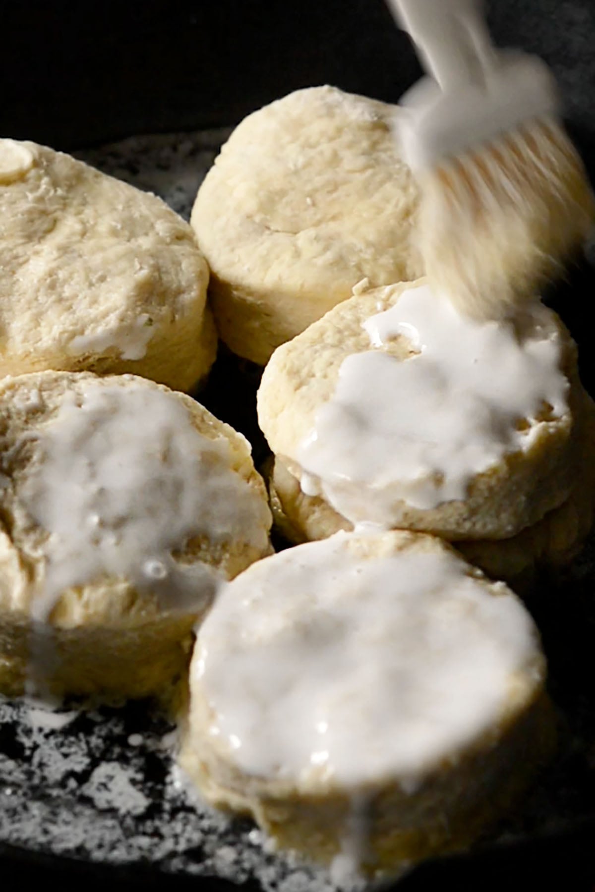 Uncooked buttermilk biscuits in a cast-iron skillet being brushed with buttermilk.