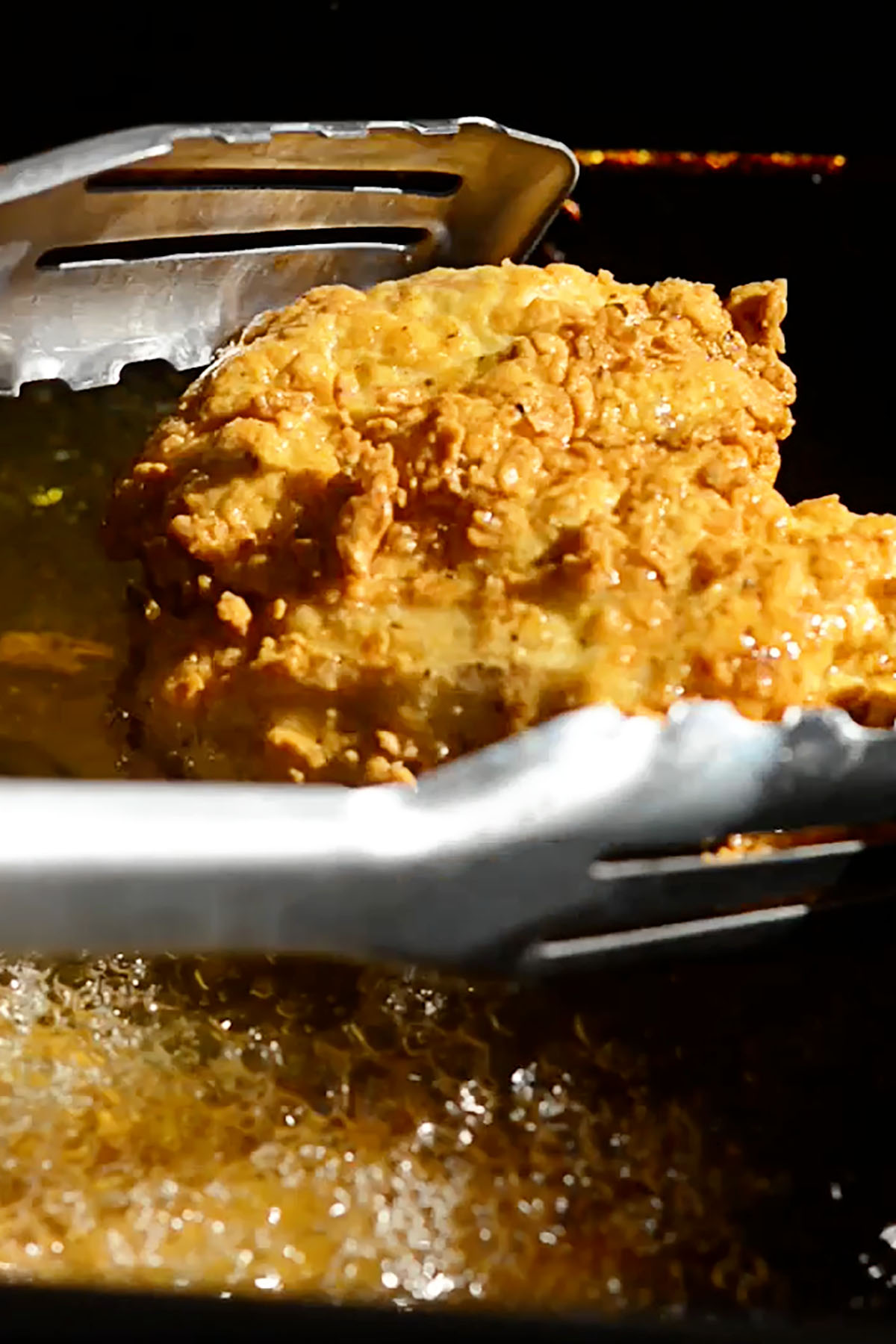 A pair of tongs holding a crunchy deep fried chicken breast over hot oil in a deep fryer.