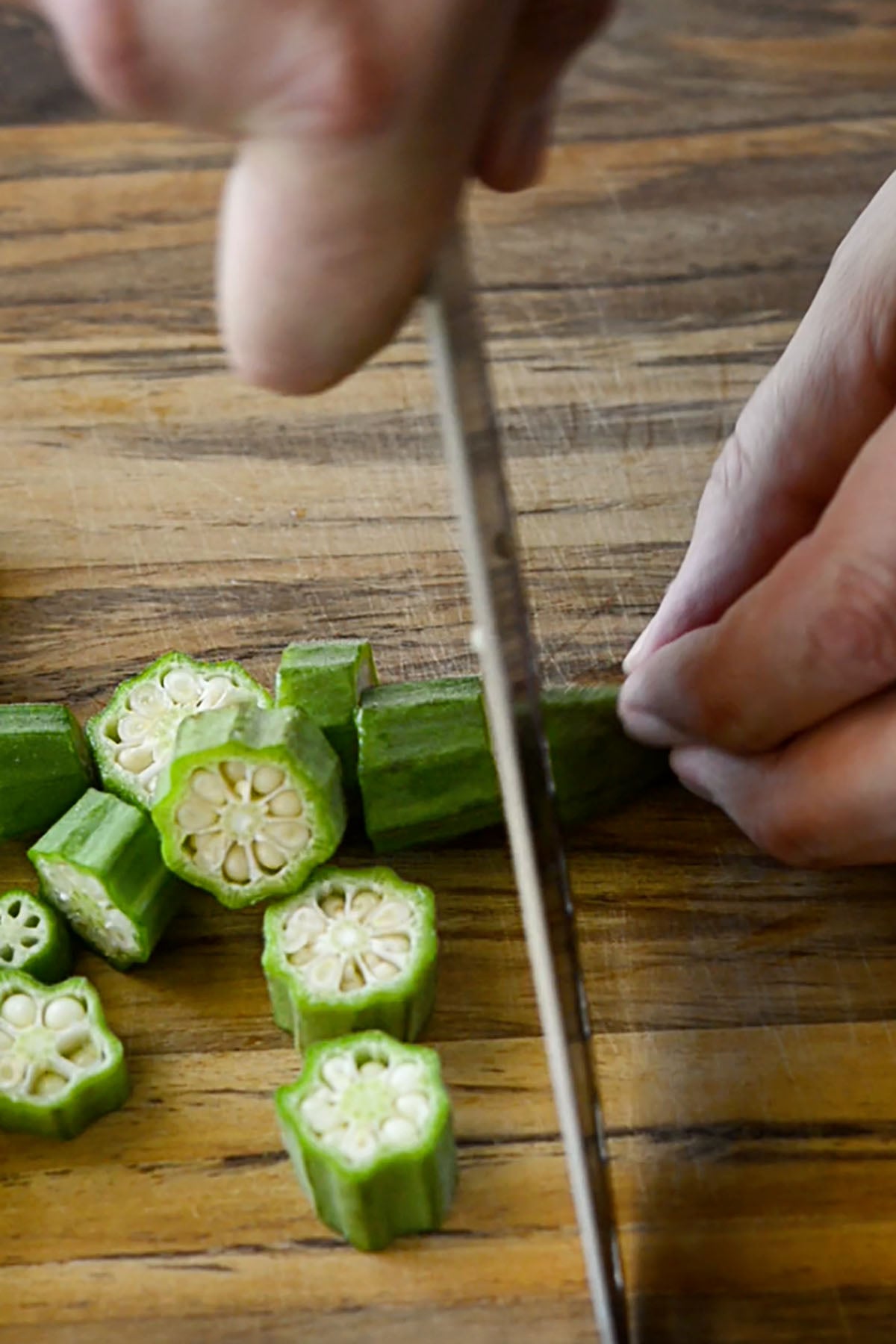 Okra being sliced into even bit sized pieces on a wood cutting board.