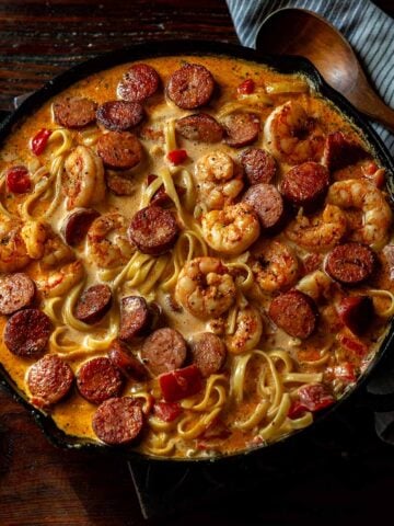 Cajun Shrimp and Sausage Pasta in a cast Iron skillet on a wooden table with a wooden spoon and towel.
