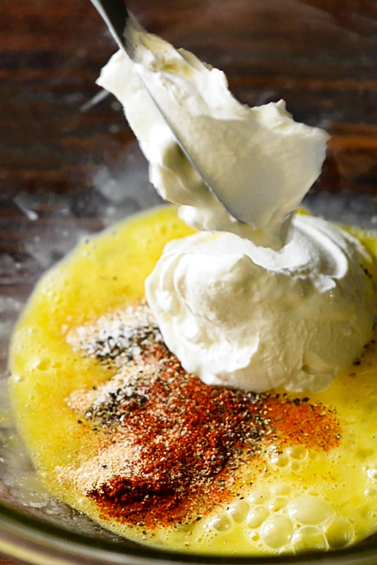 A large dollop of sour cream being added to a bowl of whisked seasoned eggs.