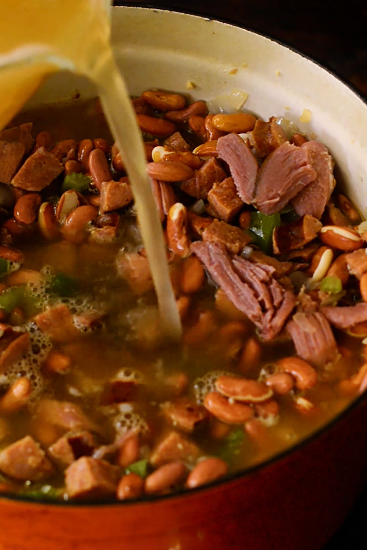 Chicken stock being poured into a dutch oven full of soaked red kidney beans, smoked turkey, cubed andouille sausage, and the holy trinity for red beans and rice.