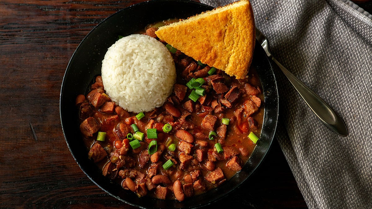 Louisiana Red Beans and Rice served fresh with a slice of cornbread and garnished with a few splashes of hot sauce and a sprinkle of freshly sliced green onions.