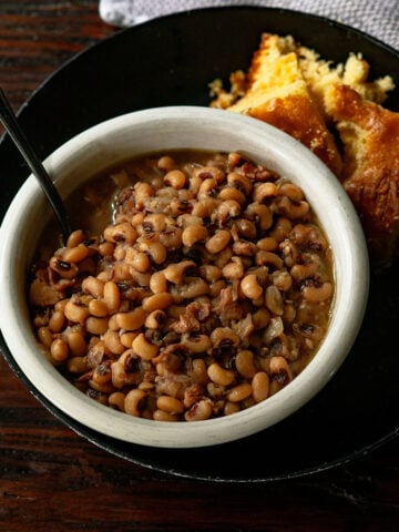 Black-Eyed Peas served in a white bowl with a black plate and fresh baked cornbread on the side.