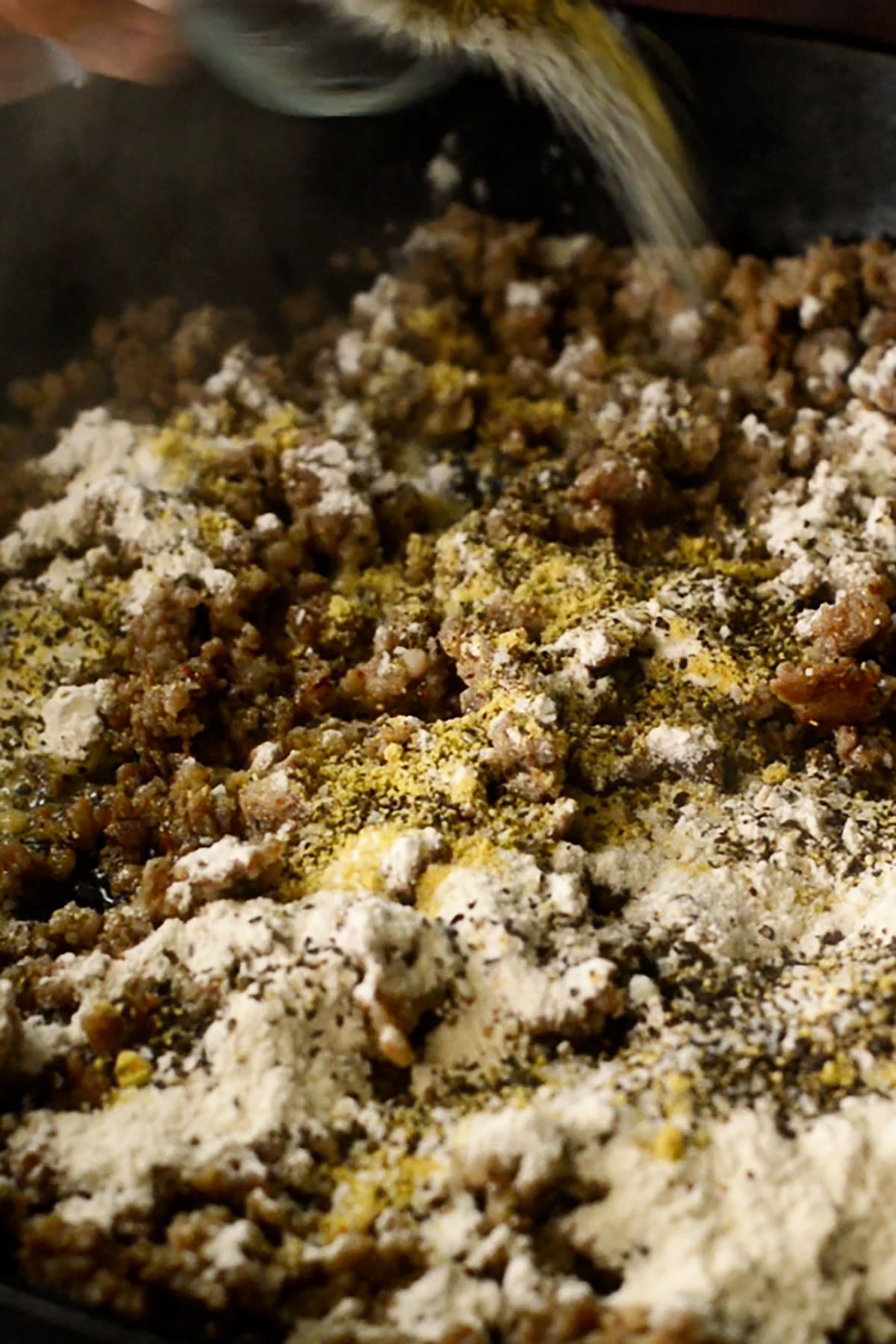 Browned breakfast sausage in a cast iron skillet topped with flour, salt, pepper, and chicken bouillon powder.