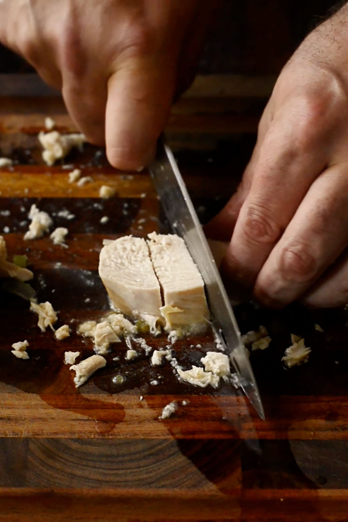 Poached chicken breast being cubed on a wood cutting board.