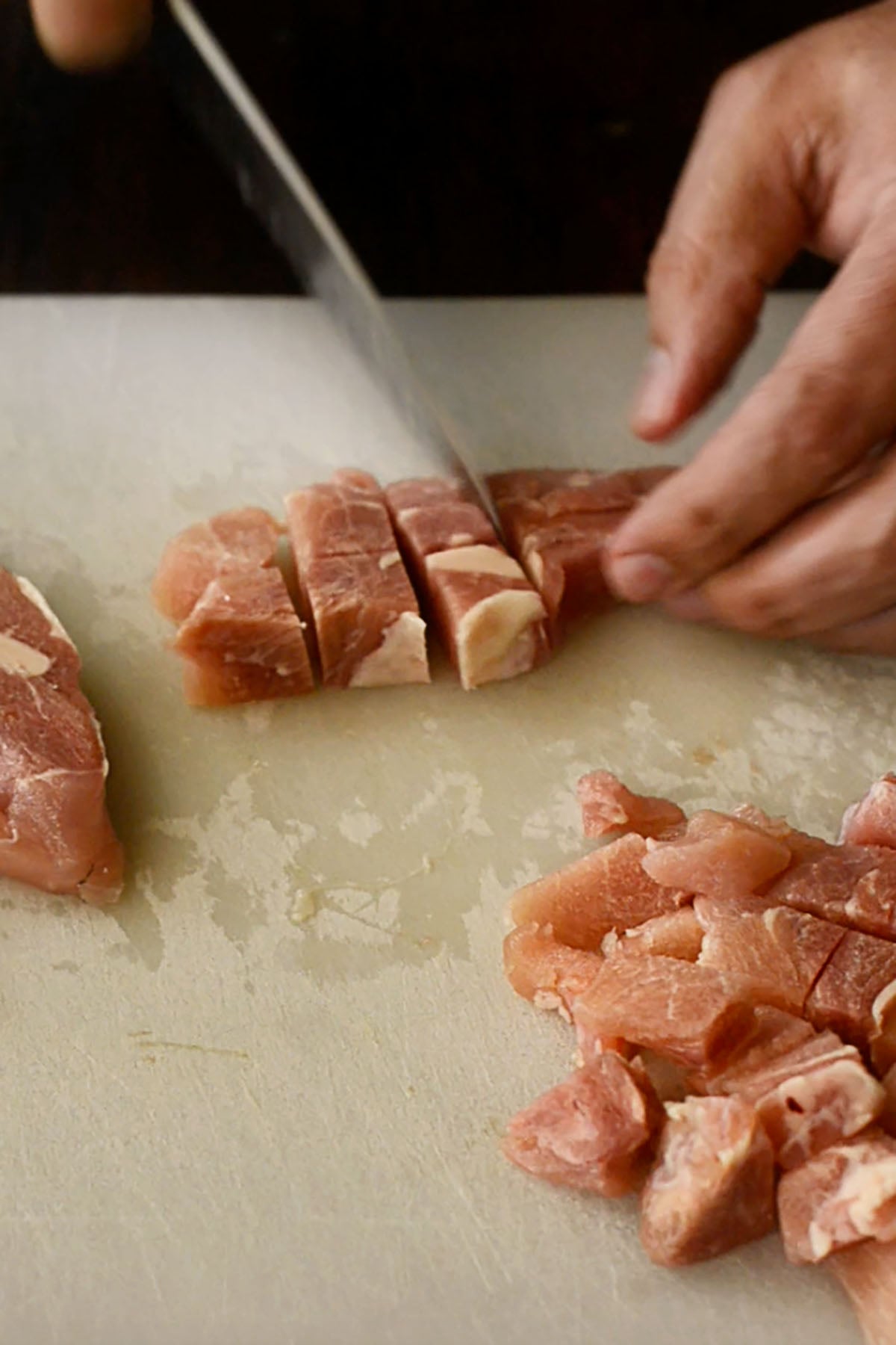 Chicken thighs being cubed on a white cutting board.