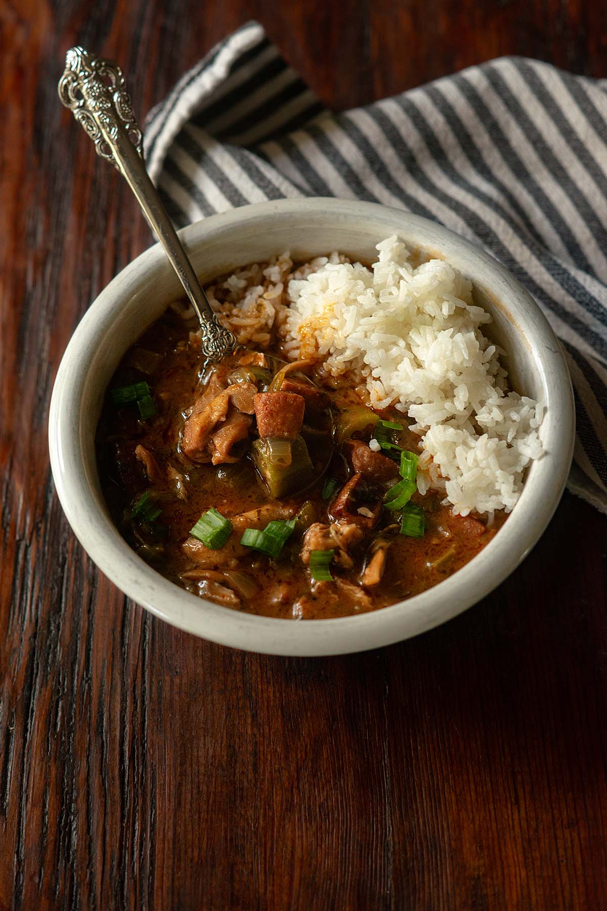 Chicken and sausage gumbo served in a white bowl with steamed rice and vintage spoon on a wooden table with a striped towel on the side.