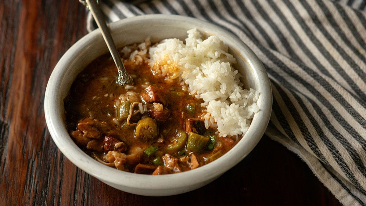 Chicken and sausage gumbo served with steamed white rice in a white bowl on a rustic wooden table.