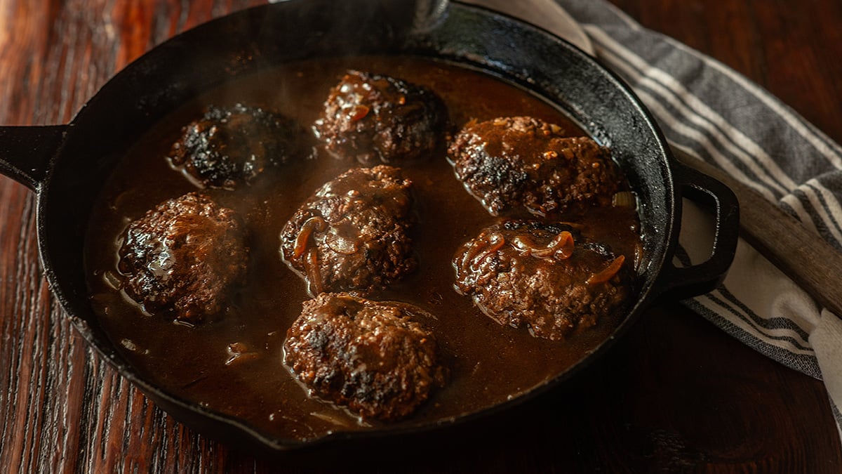 Hamburger steaks and a dark onion gravy in a cast iron skillet.